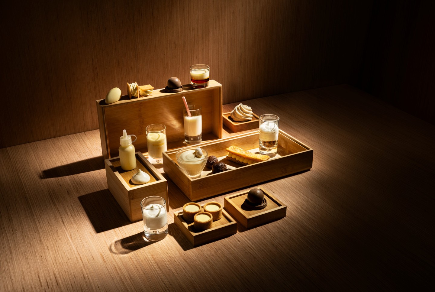 Assorted desserts and creamy drinks arranged in wooden trays on a table, illuminated by warm spotlight lighting.